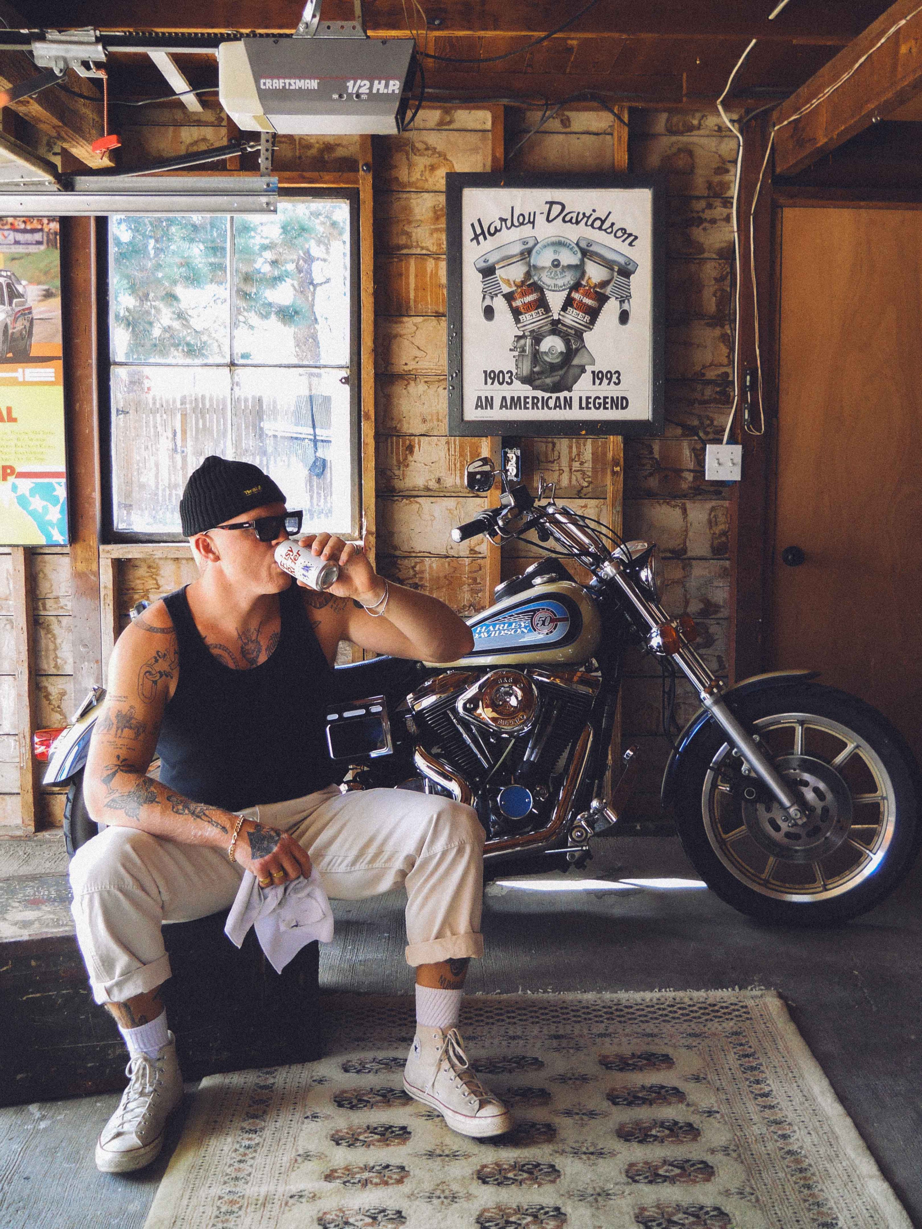 A man in drinking a beer in a garage with a motorcycle behind him