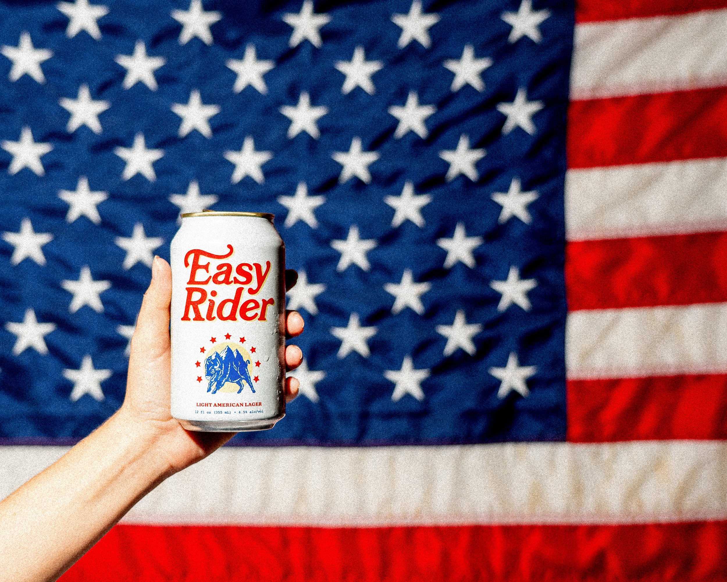 A hand holding a cold Easy Rider beer can in front of a large American flag