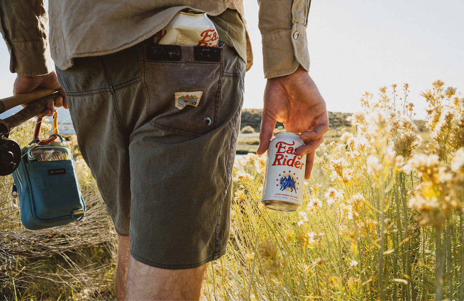 A man holding fishing gear and a can of beer walking through a field
